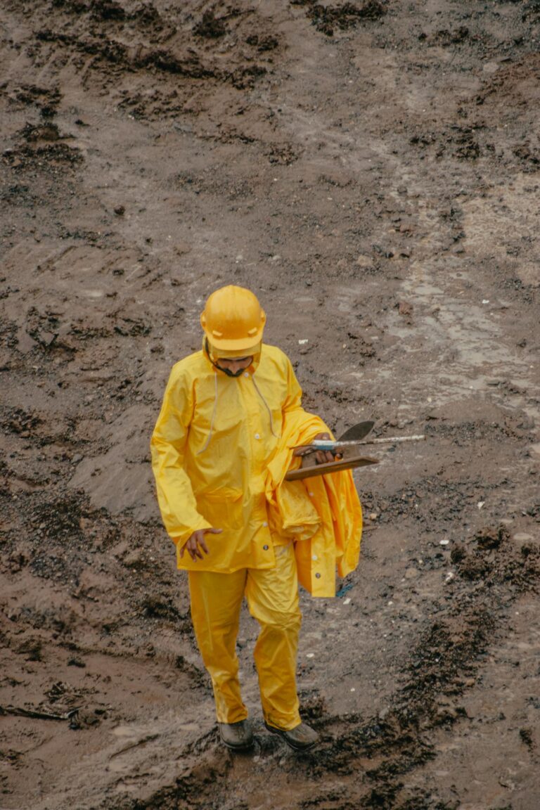 A construction worker in yellow rain gear and helmet holding a clipboard stands on a muddy site.