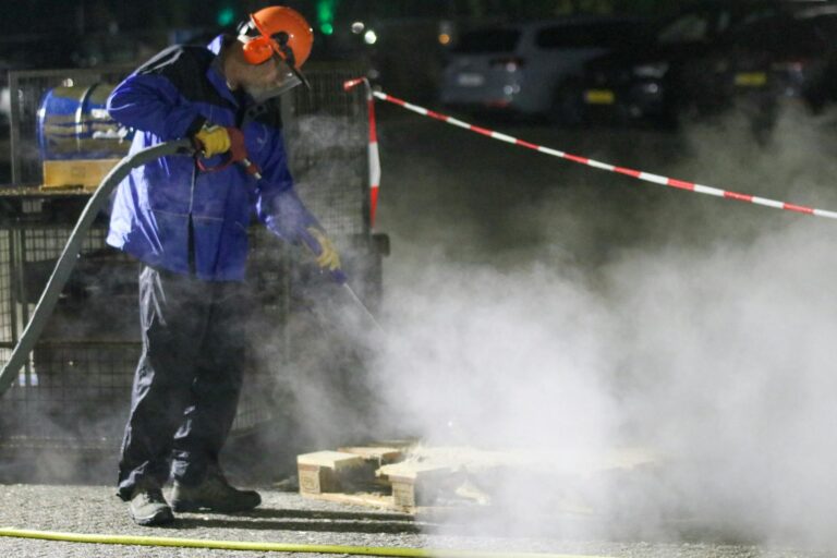 Man in protective gear using a high-pressure washer amid steam at night in an industrial setting.